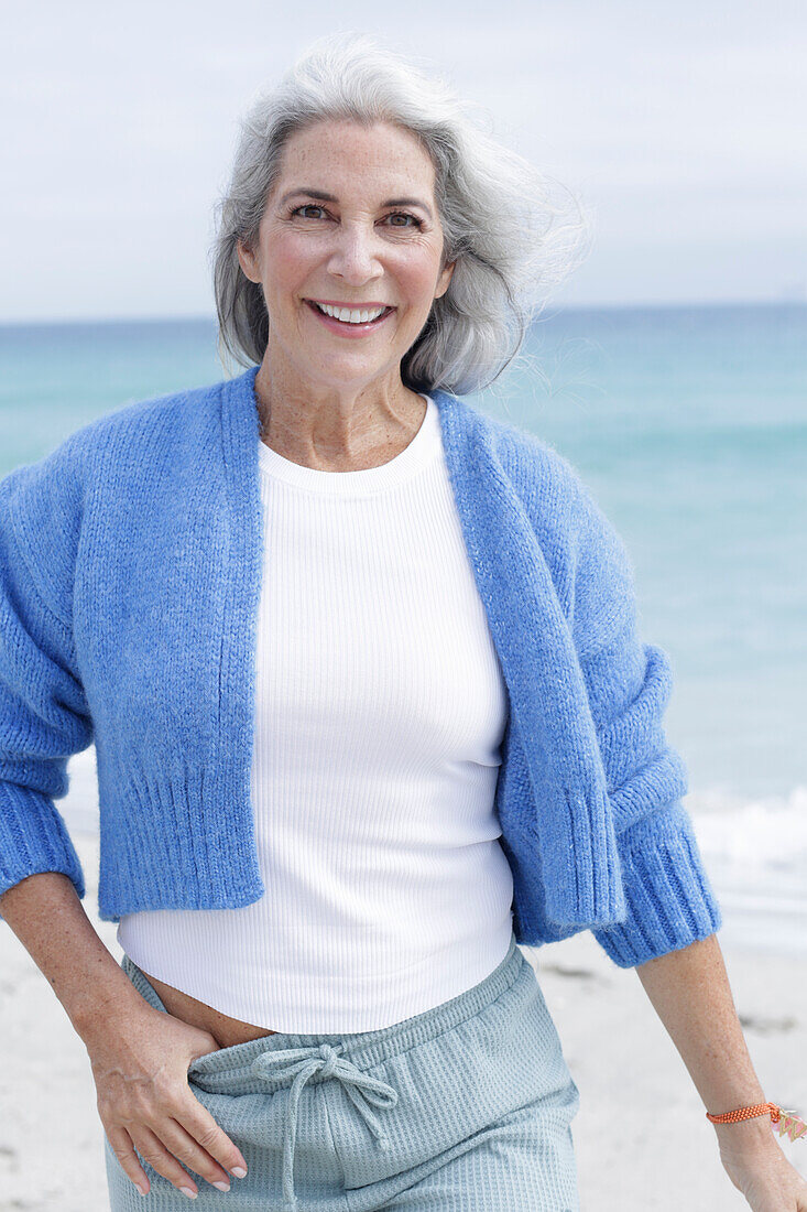 Mature woman with grey hair in white t-shirt, blue cardigan and trousers on the beach