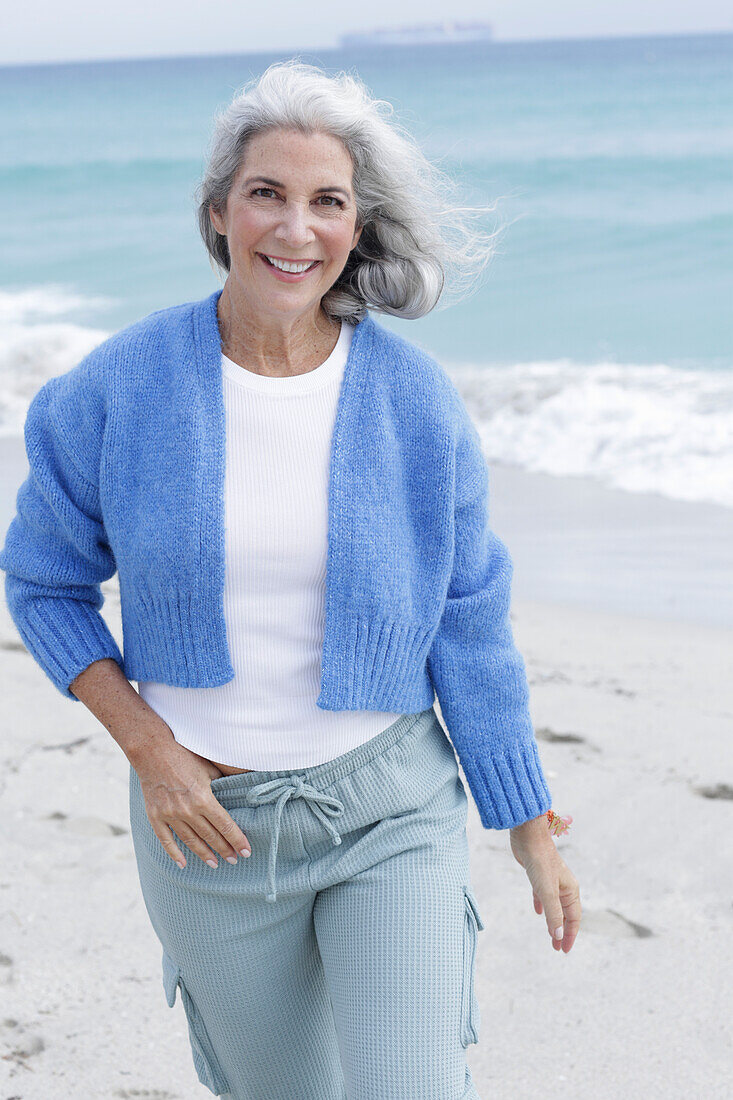 Mature woman with grey hair in white t-shirt, blue cardigan and trousers on the beach