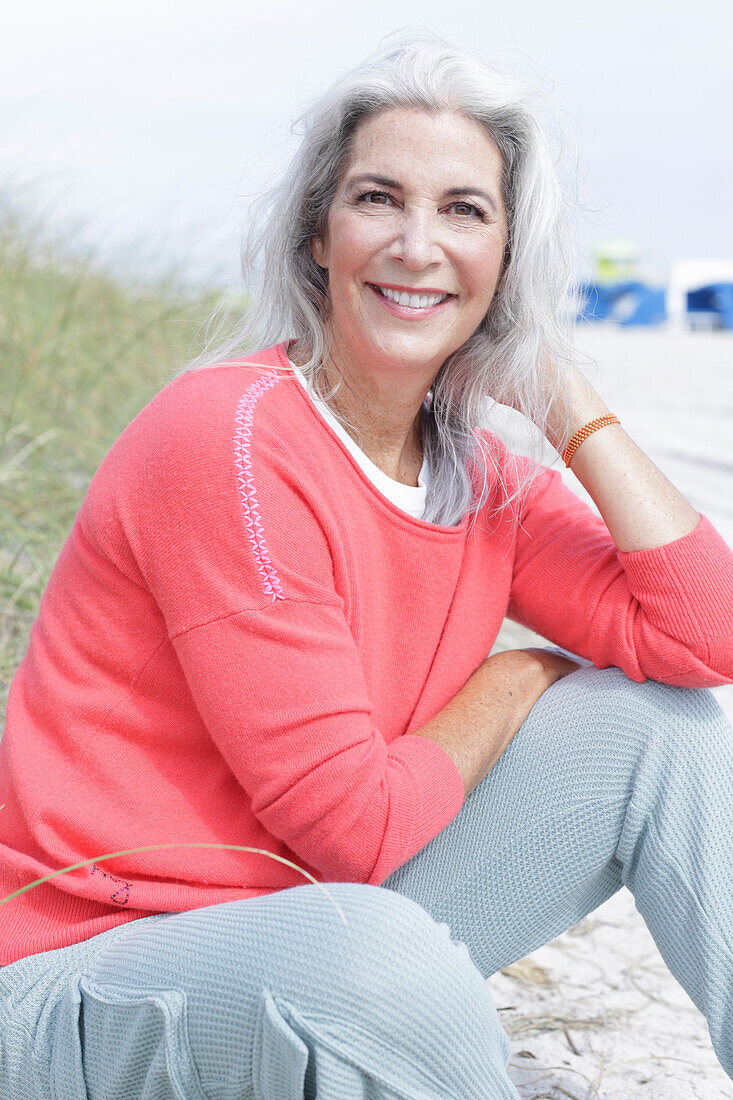 Reife Frau mit grauen Haaren in lachsfarbenem Pullover und Hose am Strand