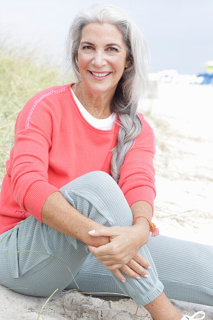 Reife Frau mit grauen Haaren in lachsfarbenem Pullover und Hose am Strand