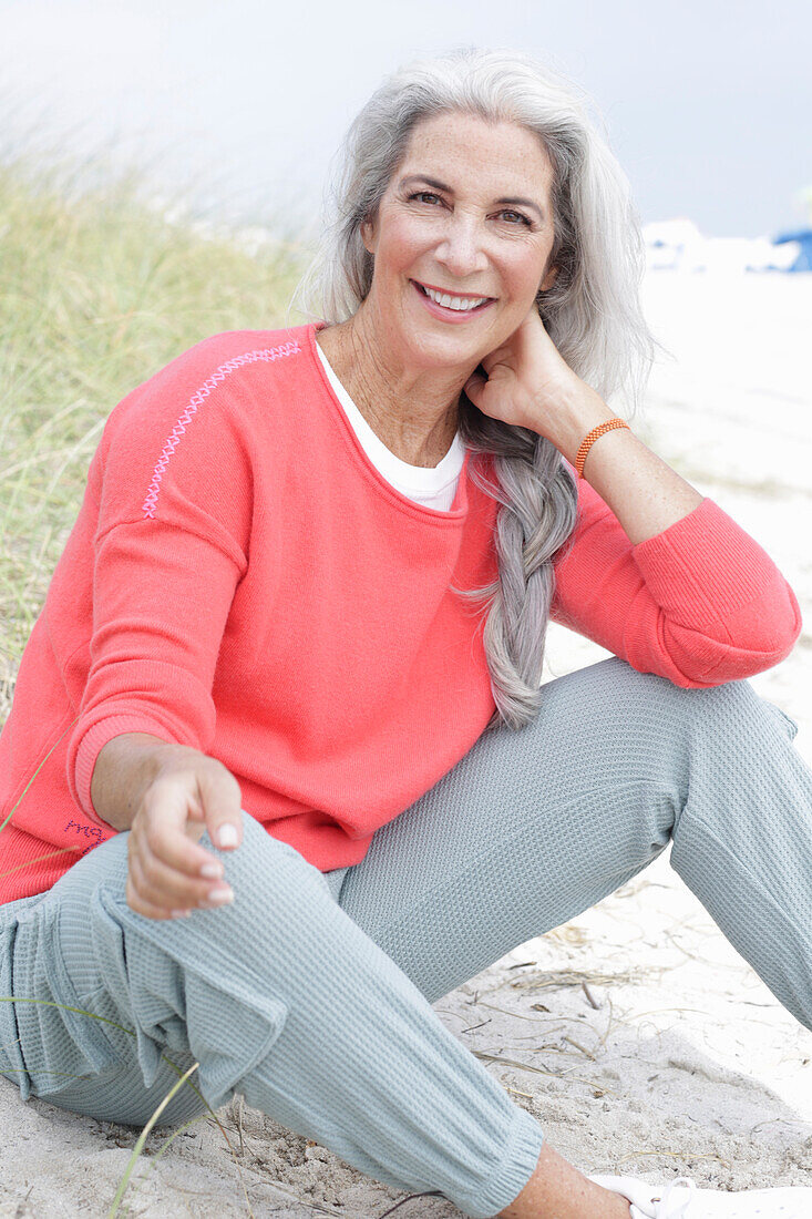 Reife Frau mit grauen Haaren in lachsfarbenem Pullover und Hose am Strand