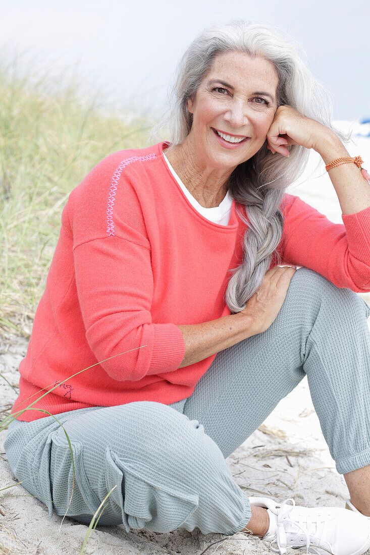 Reife Frau mit grauen Haaren in lachsfarbenem Pullover und Hose am Strand