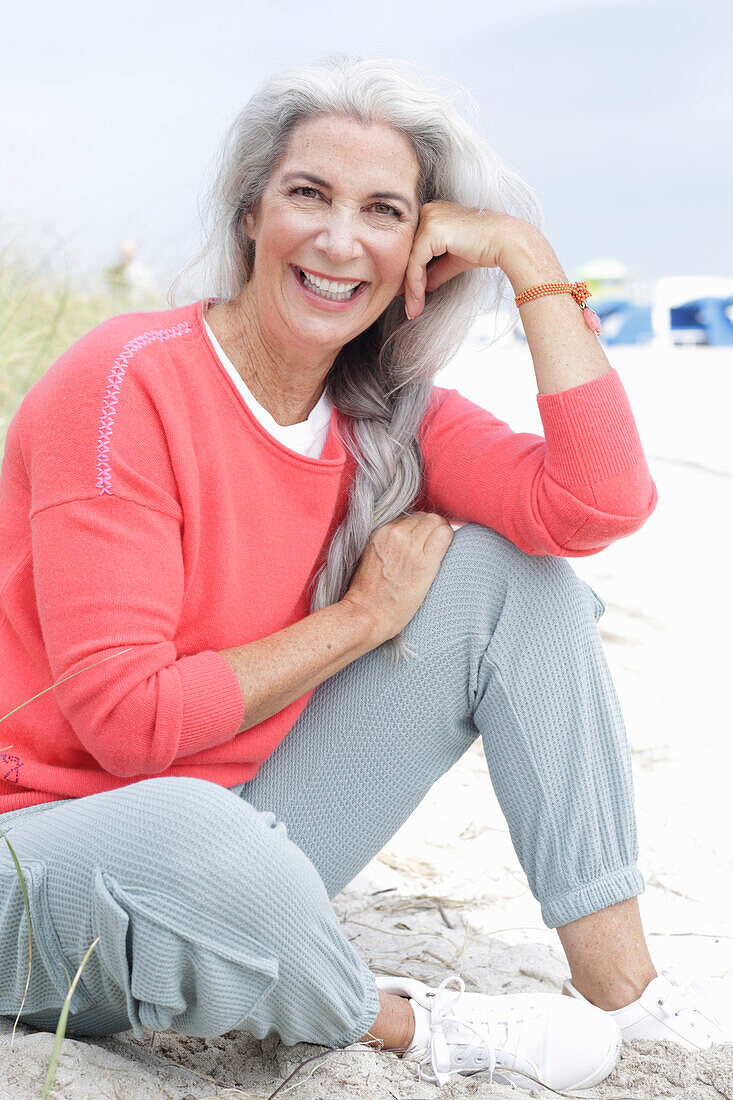 Reife Frau mit grauen Haaren in lachsfarbenem Pullover und Hose am Strand