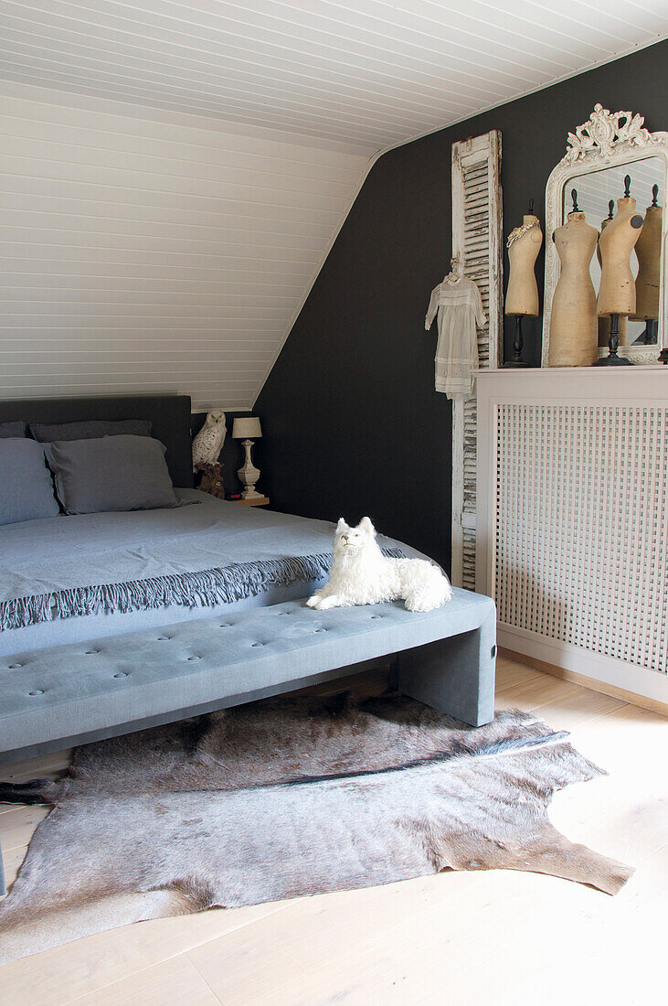 Double bed with grey-blue bedspread, matching clothes bench with stuffed animal, dressmaker's dummies on top of radiator cover