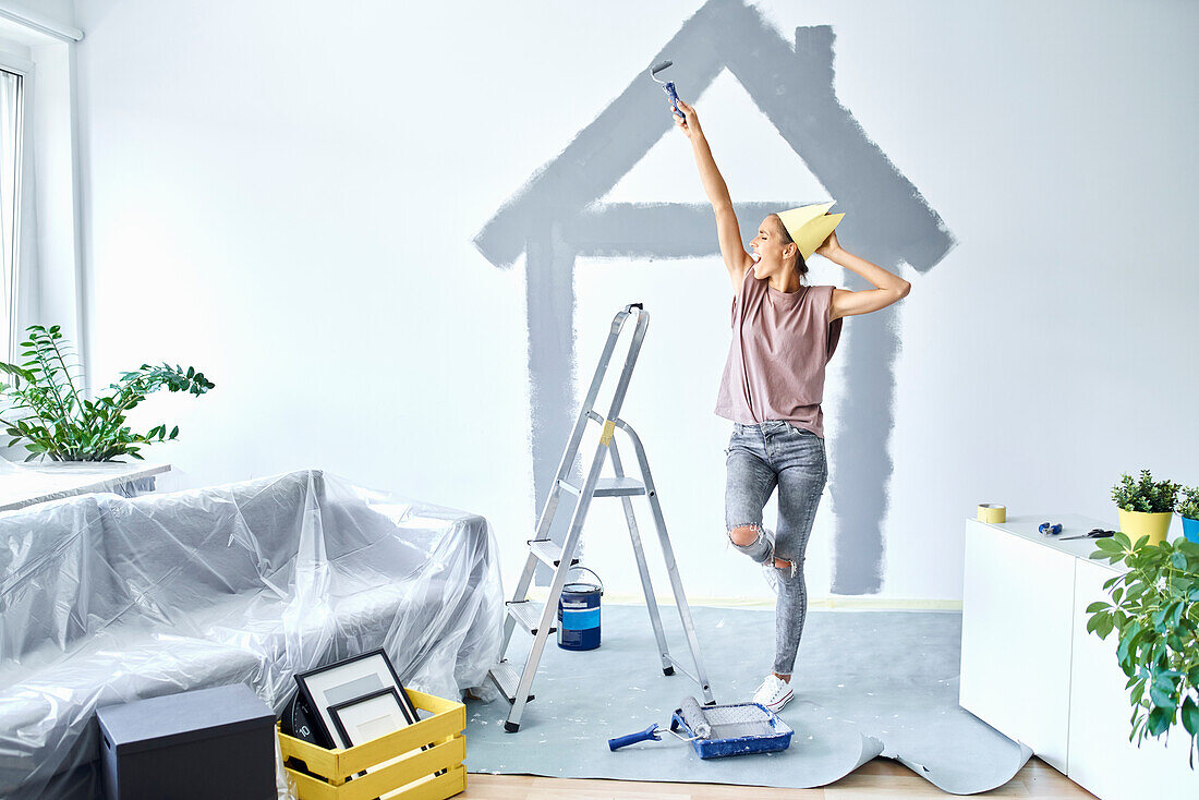 Glückliche junge Frau mit Papierkrone, die mit erhobener Hand gegen das Haus steht, das zu Hause an die Wand gemalt wurde