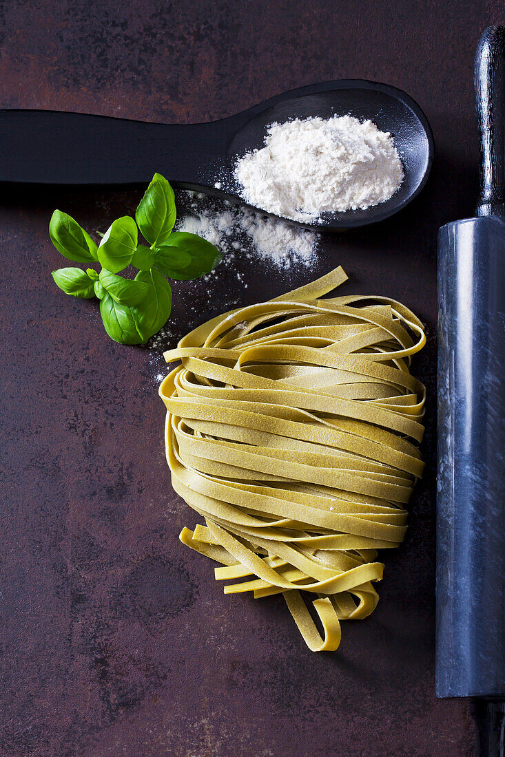 Green Tagliatelle, spoon of flour, basil leaves and rolling pin on rusty ground