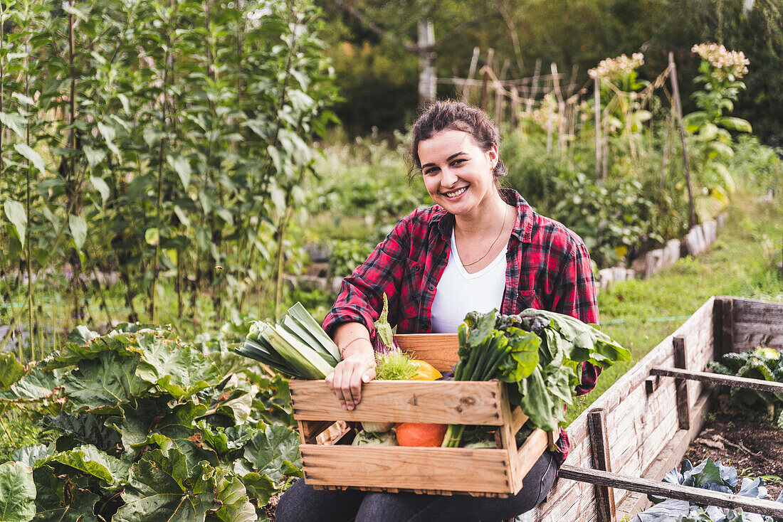 Lächelnde junge Frau mit Gemüse in Kiste, die gegen Pflanzen im Garten sitzt
