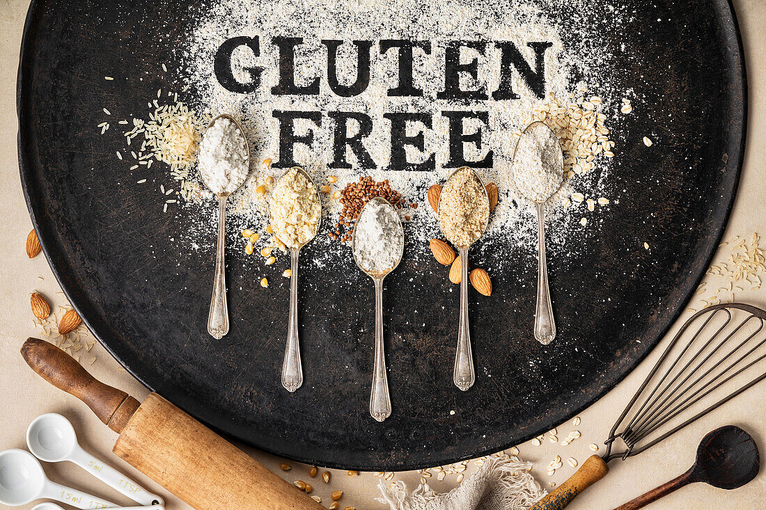 'Gluten free' Schriftzug in Mehl auf Vintage-Backblech und Löffel mit verschiedenen glutenfreien Mehlsorten
