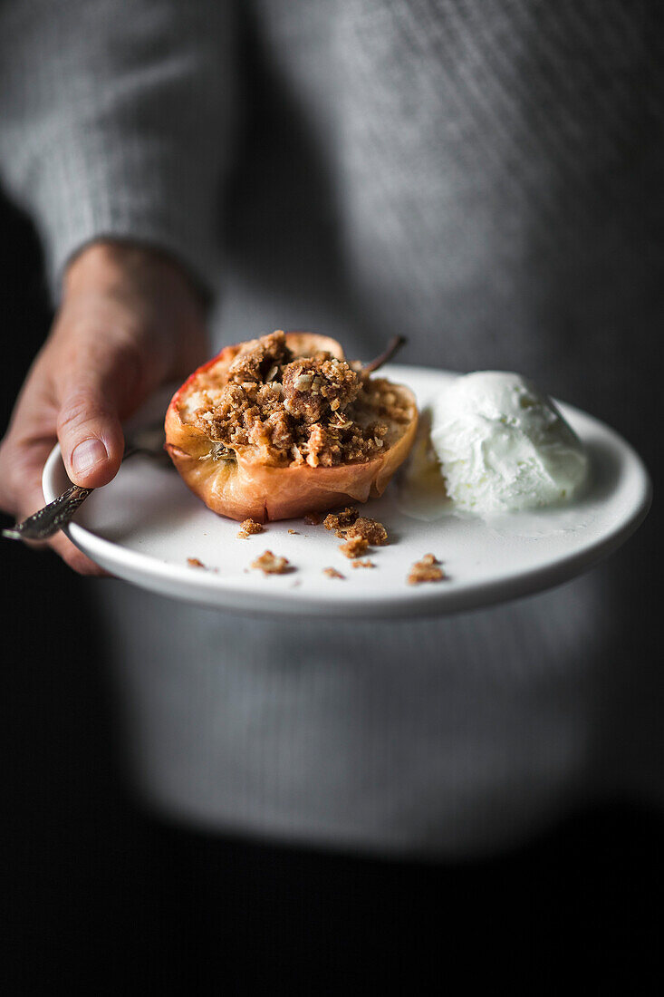 A baker holding a plate of baked apple with streusel and ice cream