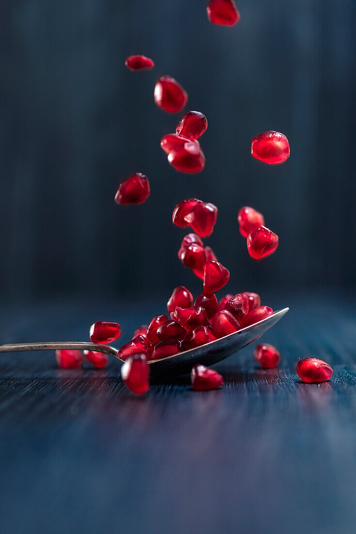 Red fresh juicy pomegranate fruits seeds falling on spoon and dark wooden surface on blue background