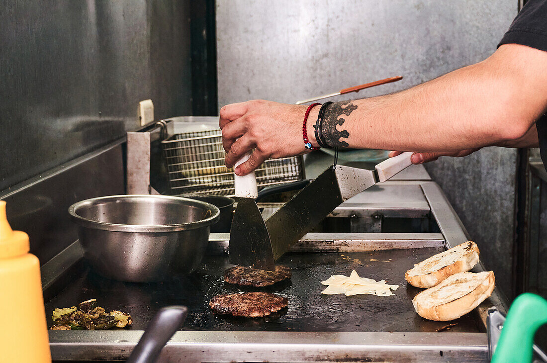 Side view of crop unrecognizable cook frying and turning over burger cutlets with spatulas in a food truck