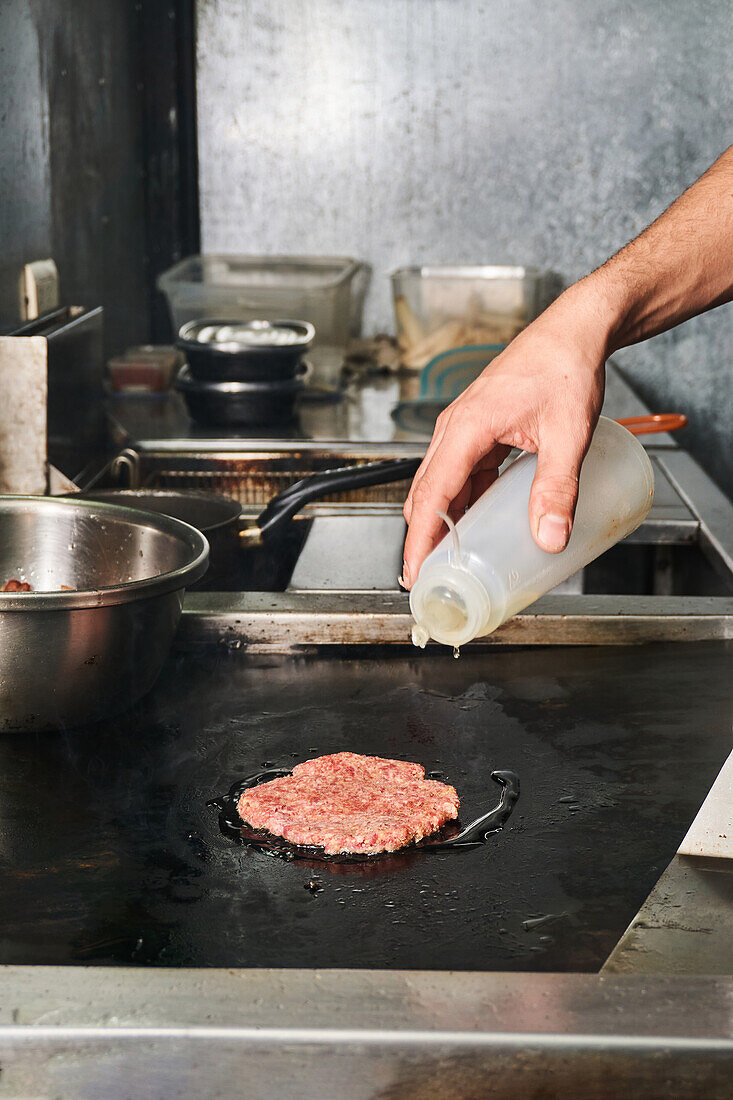 Unrecognizable chef pouring vegetable oil from bottle on raw patty while cooking burger in restaurant kitchen