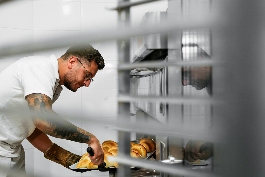 Male baker with tattoos on arm baking croissants in large metal oven during work in bakery