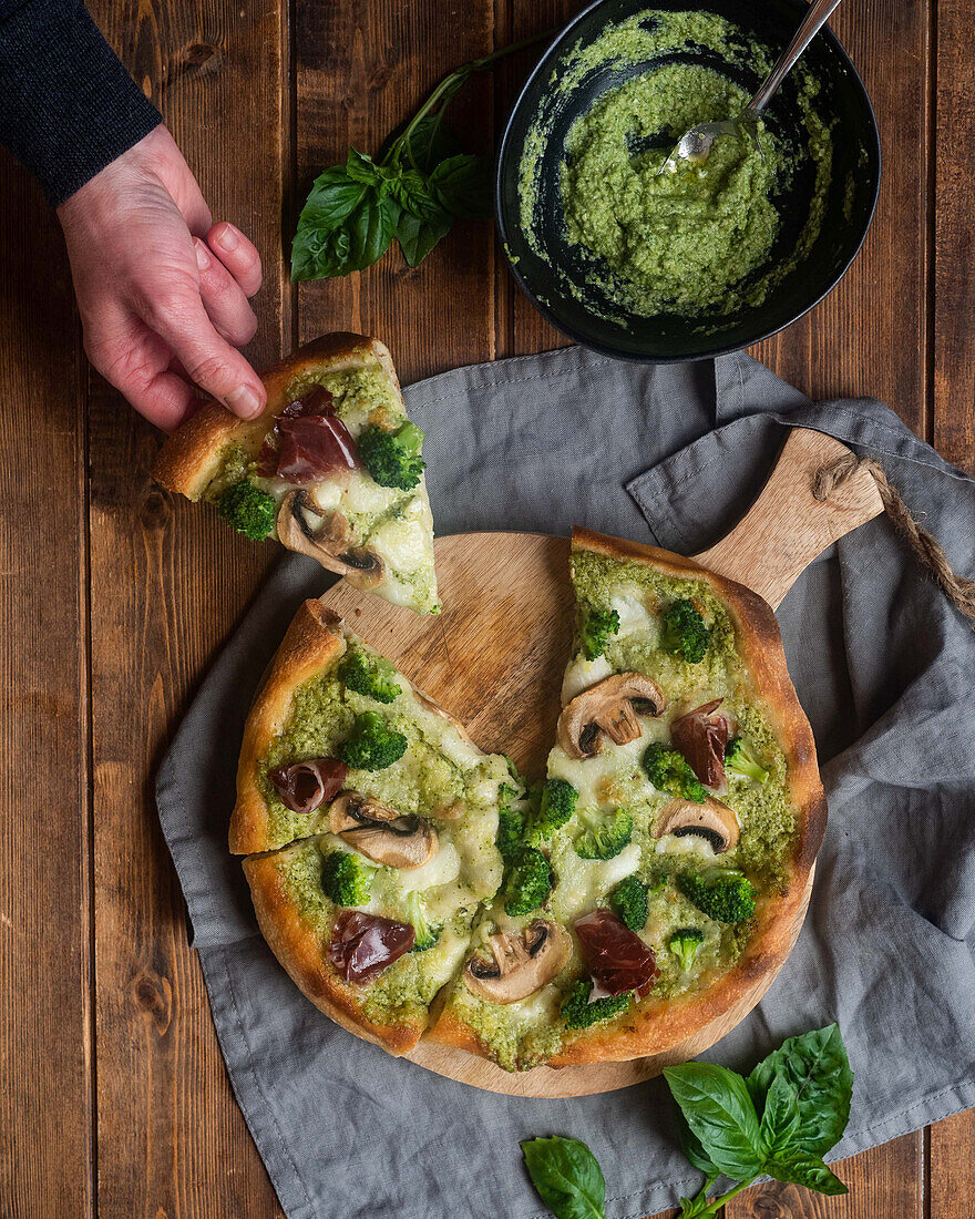 Unrecognizable person taking slice of tasty pizza with pesto sauce and mushrooms served on wooden cutting board on table