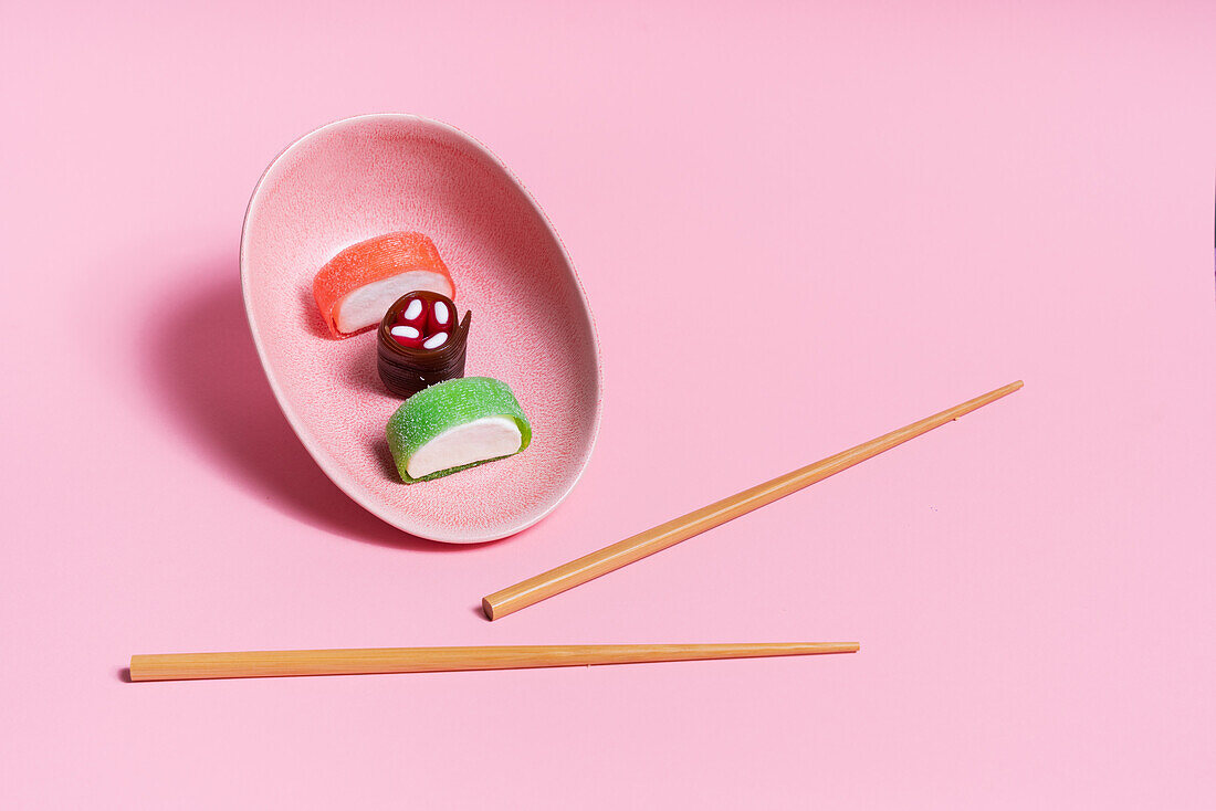 Colorful sweet sushi served on ceramic plate with wooden chopsticks placed on pink background