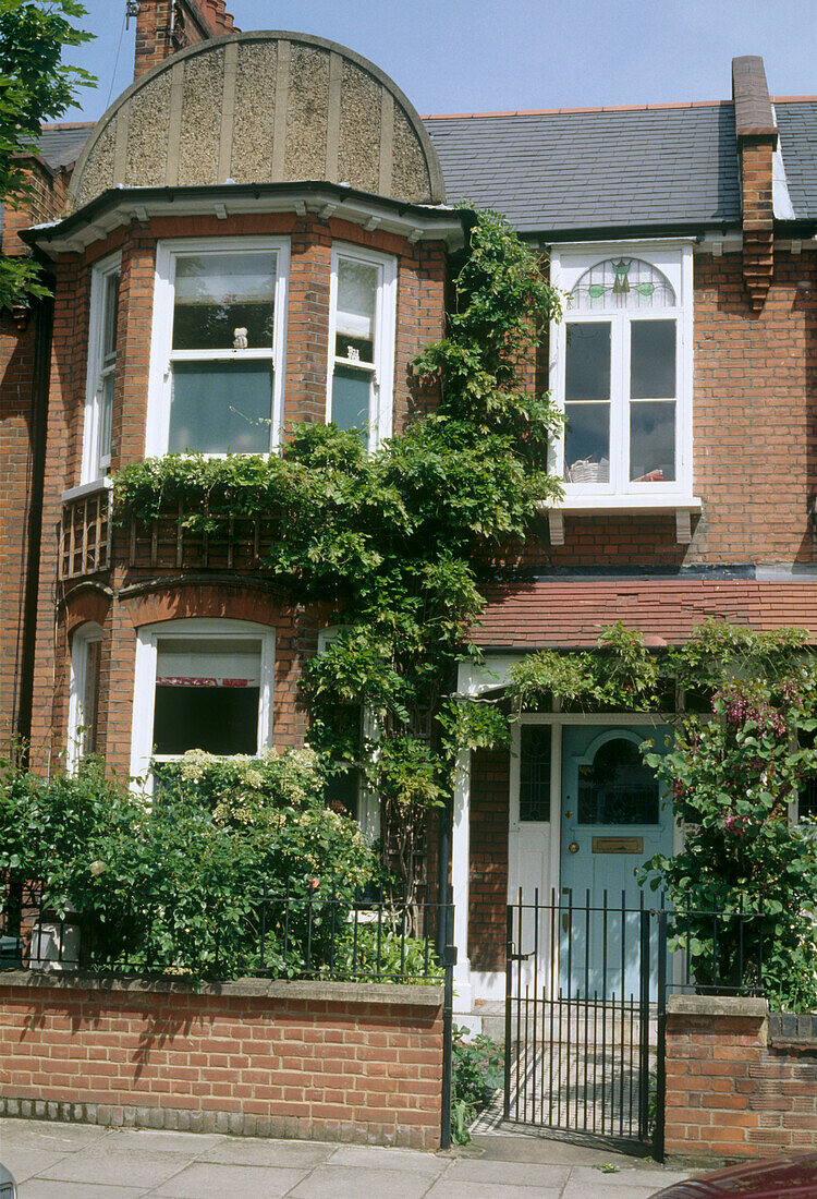 The exterior of a traditional pre war semi detached house covered in plants with a front garden and black metal gate