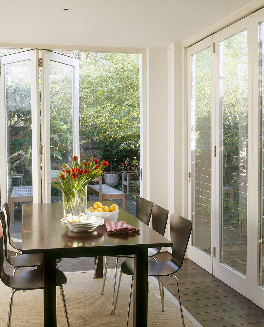 A conservatory dining room with glass walls surrounding a modern dining table and chairs laden with food and flowers