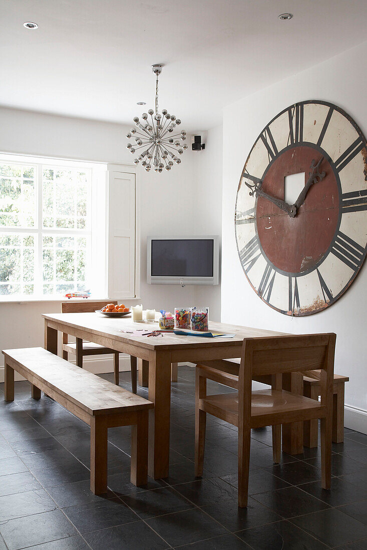 Oversize clock on wall next to television in dining room with wooden table and chairs