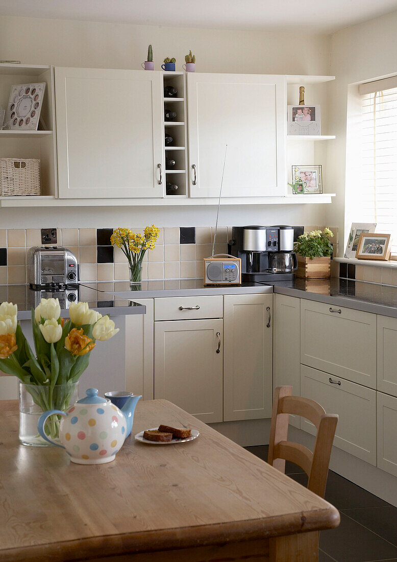 Eine moderne Küche im Landhausstil mit weißen Schränken, einem Frühstückstisch aus Holz und Tulpen in einer Vase
