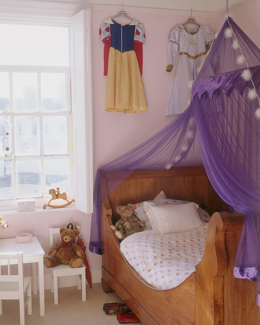 Ein traditionelles Kinderzimmer mit Holzbett und lila Betthimmel, darüber hängen zwei Kleidern an rosafarben Wand