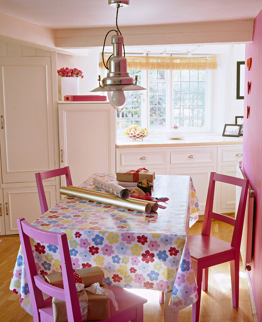 Essecke in Landhausküche mit rosa gestrichenen Stühlen, Tischdecke mit Blumenmuster, Geschenkpapierollen auf dem Tisch und Hängelampe