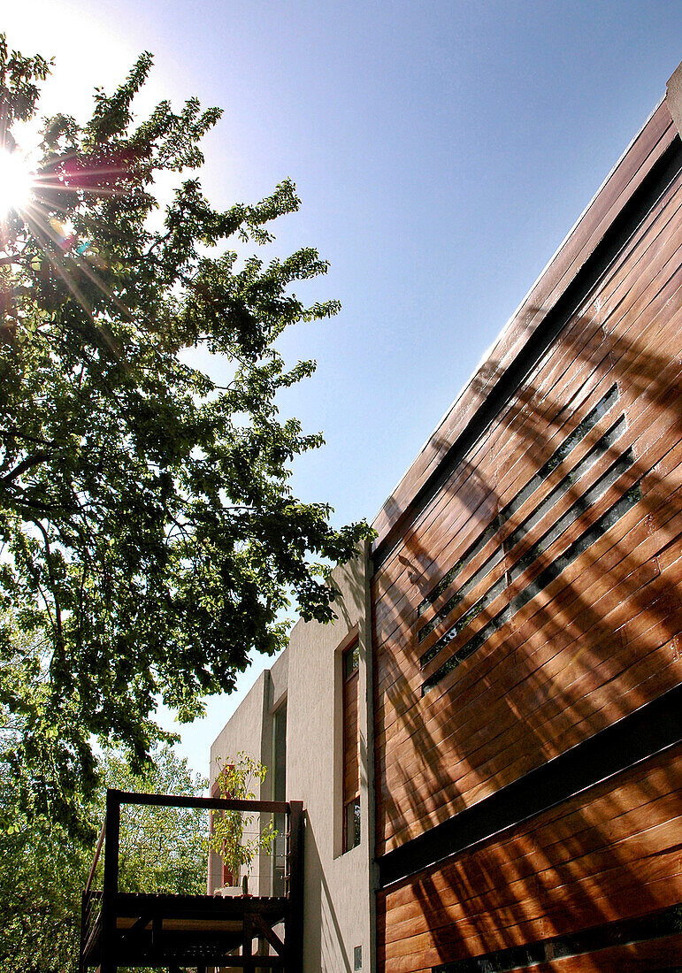 Die Fassade zeigt eine Struktur aus rostig anmutendem Eisen, die dem Eingang Schatten spendet und im Obergeschoss in einen terrassenförmigen Balkon übergeht