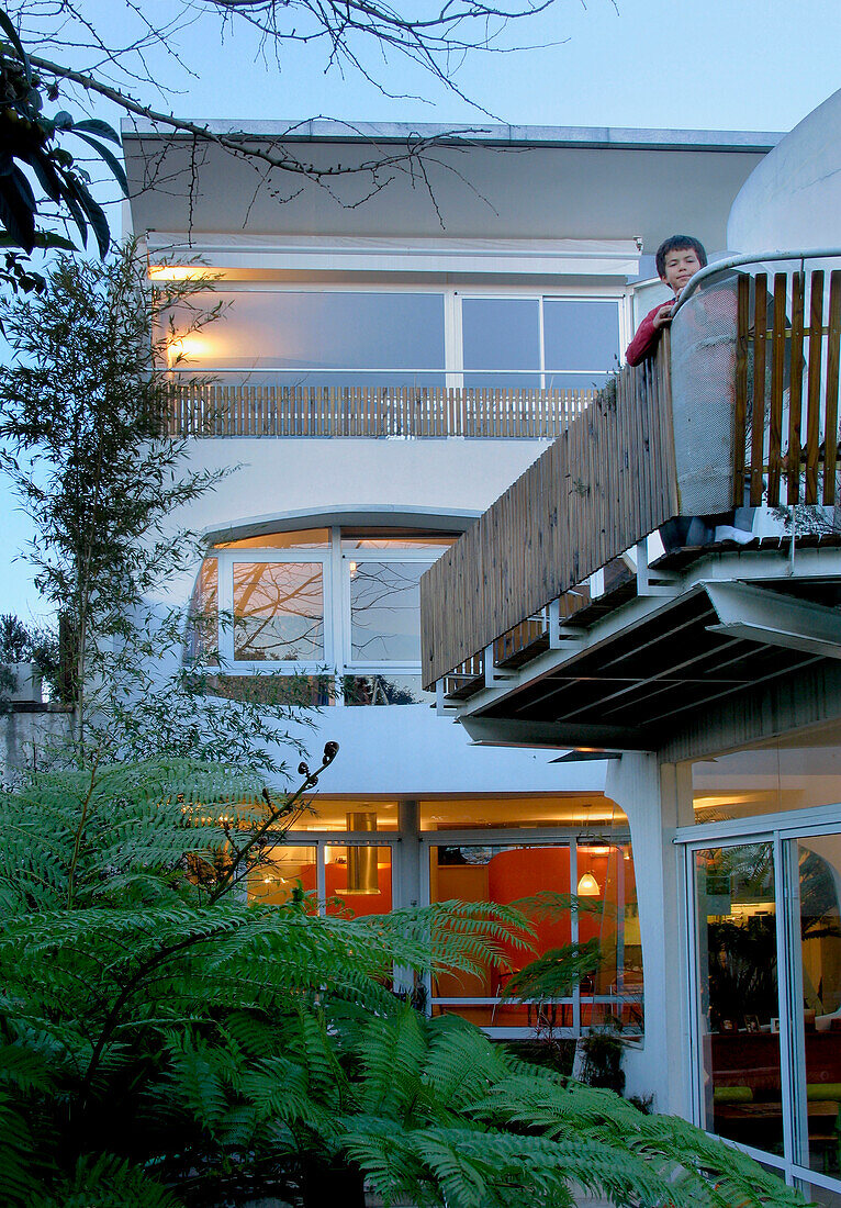 Boy standing on balcony of stucco finished white aluminium house above ferns