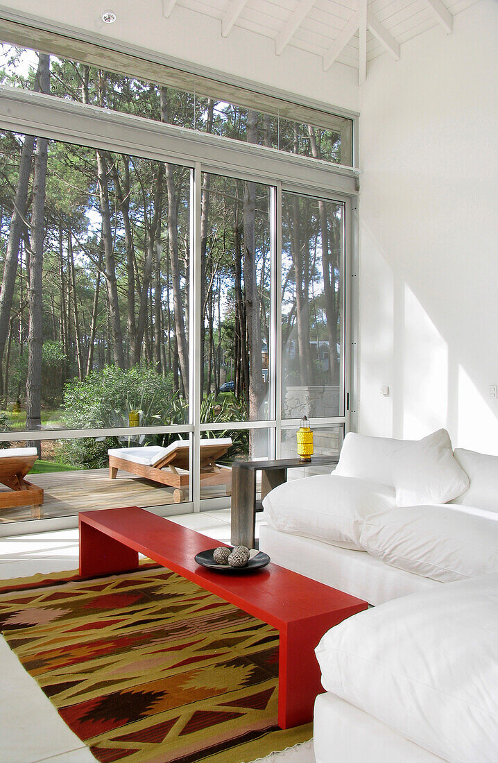 Modern bright white living room with rug and red coffee table with large windows overlooking woodland garden and terrace