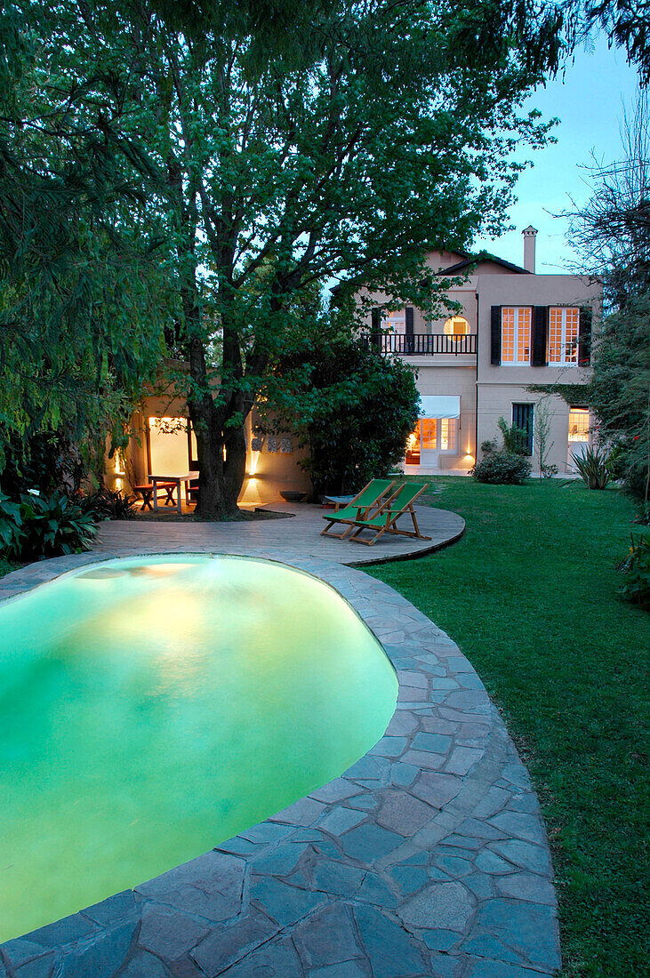 Illuminated pool with green deck chairs under tree and early 20th century building exterior