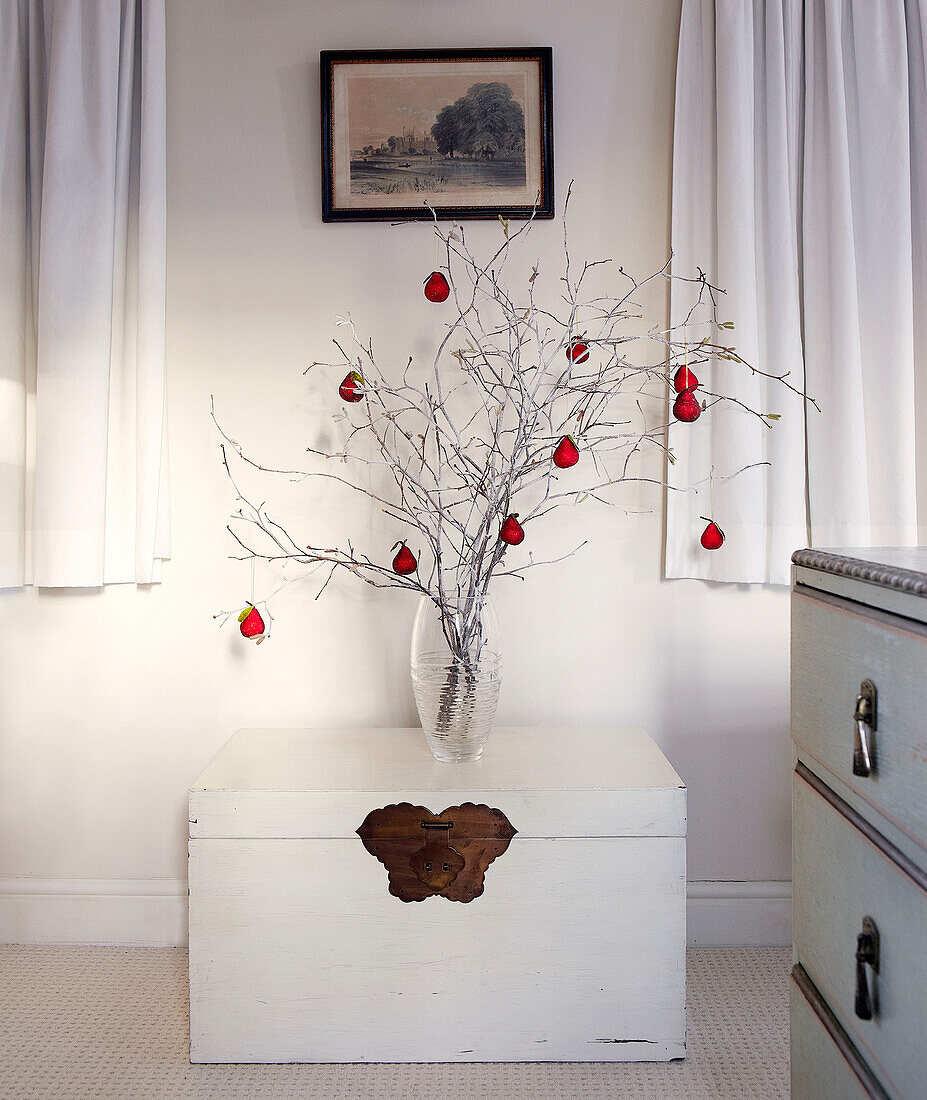 Red Christmas baubles hang on twig arrangement in glass vase on traveling chest