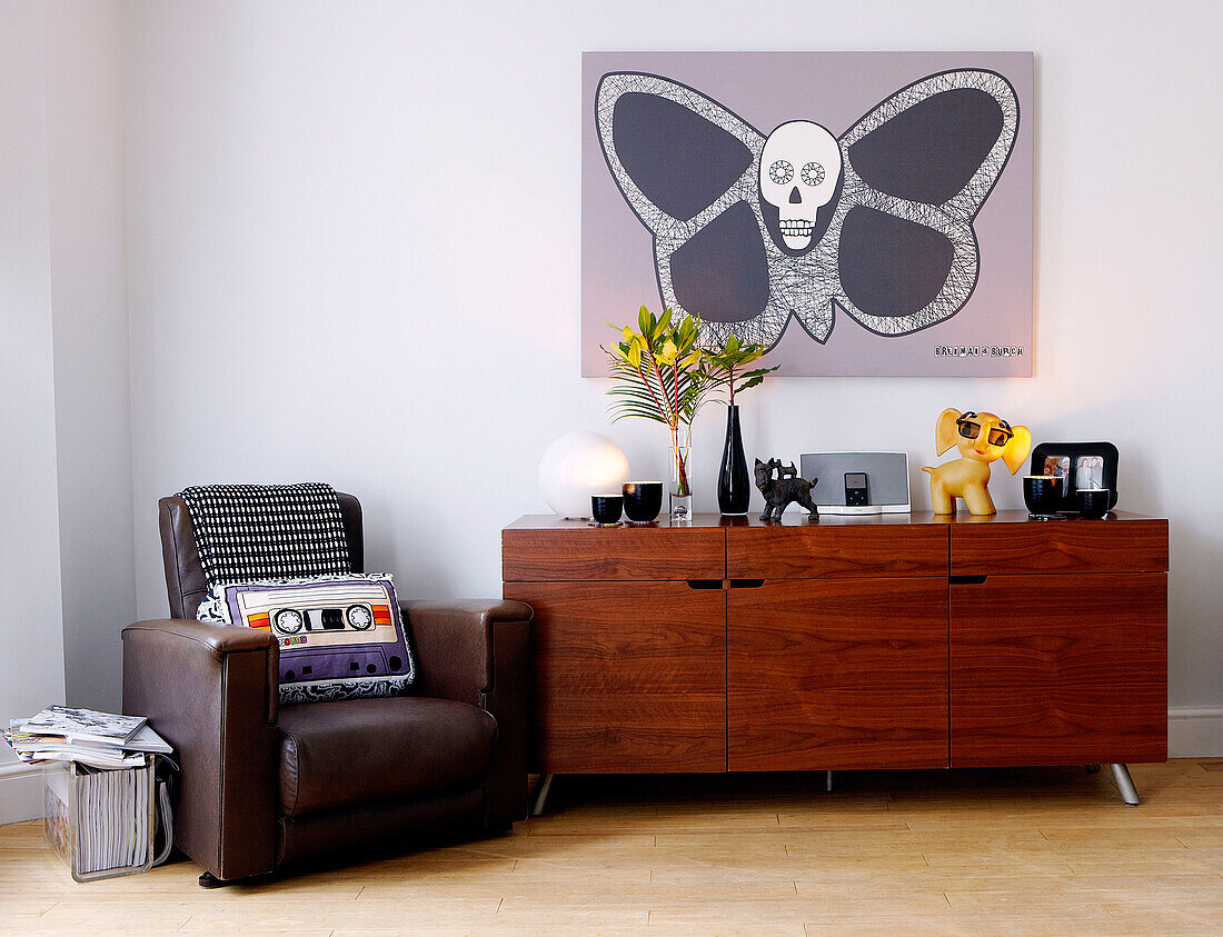 Brown leather armchair and polished wooden sideboard below unusual artwork with butterfly and skull
