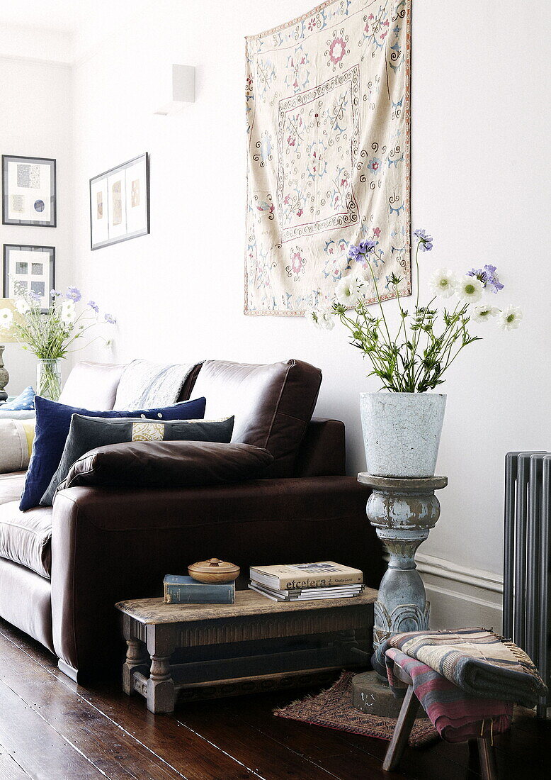 Flower arrangement on vintage plinth with brown leather sofa and wall hanging in living room of London home UK
