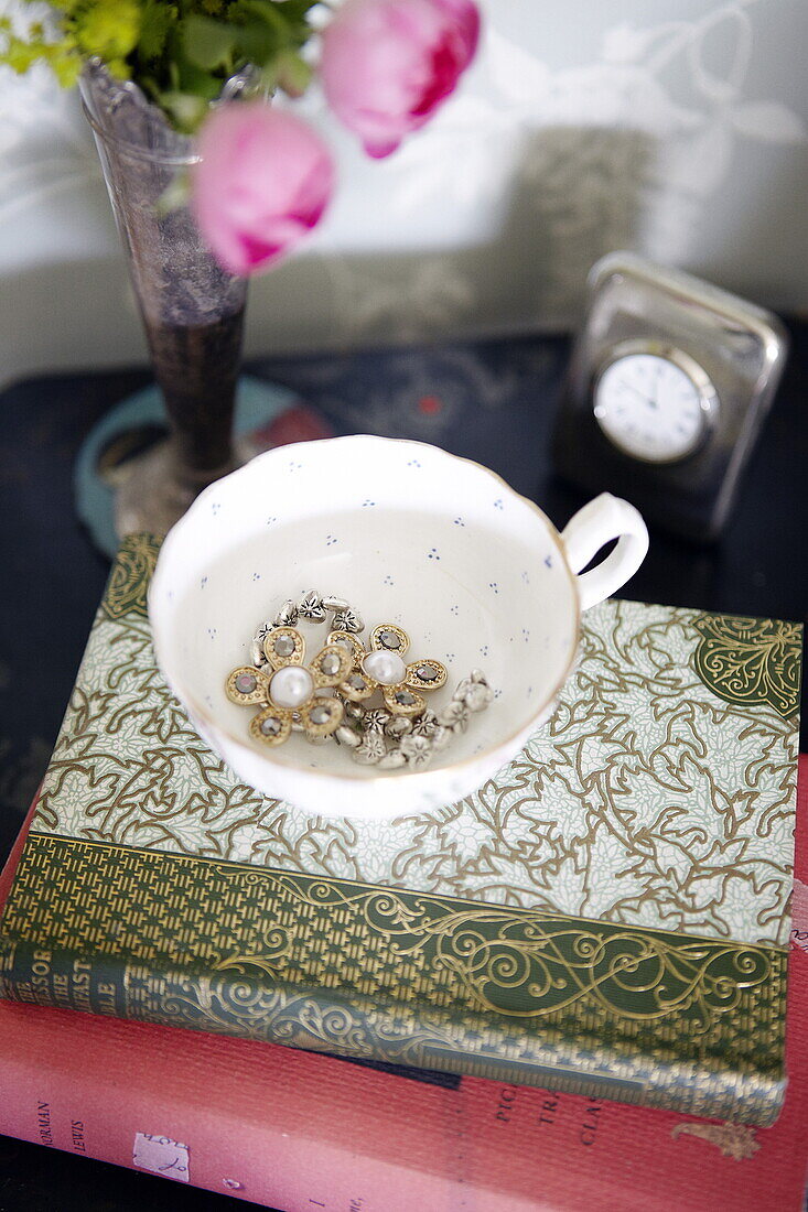 Schmuck in einer Teetasse mit gebundenen Büchern auf dem Nachttisch in einem Haus in Oxfordshire, England, UK