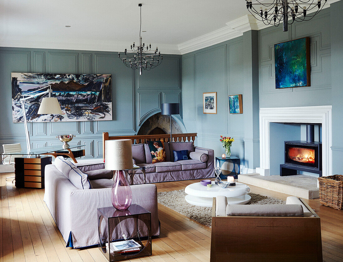 Lit fire and modern artwork with lilac sofas in panelled living room of Northumbrian manor house England UK