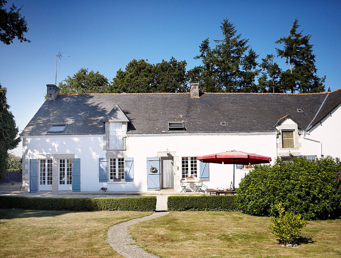 Roter Sonnenschirm auf der Terrasse eines weiß getünchten bretonischen Bauernhauses mit blauen Fensterläden Frankreich