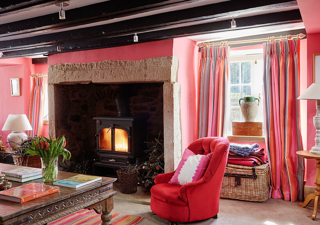 Red armchair at lit fireside in pink living room of 18th century Northumbrian mill house, UK