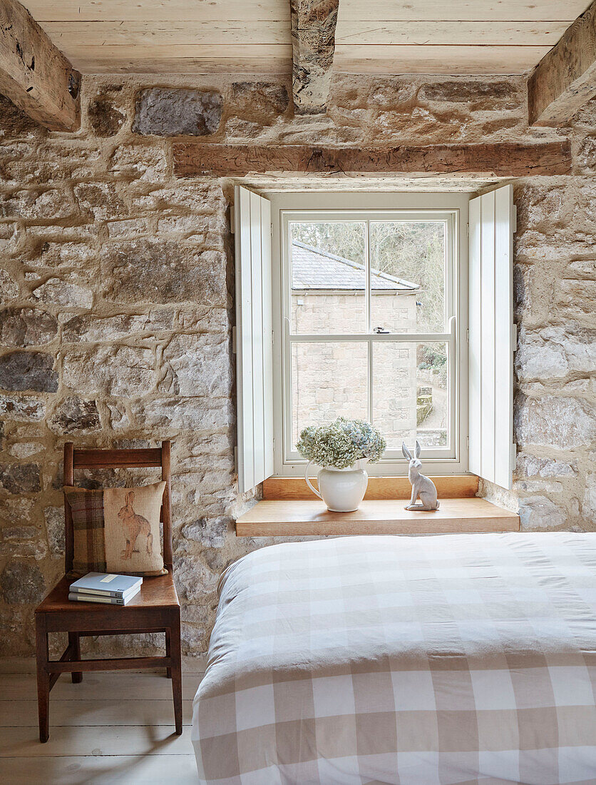 Wooden chair beside window with shutters in exposed stone bedroom of Grade II listed Tudor bastle Northumberland UK