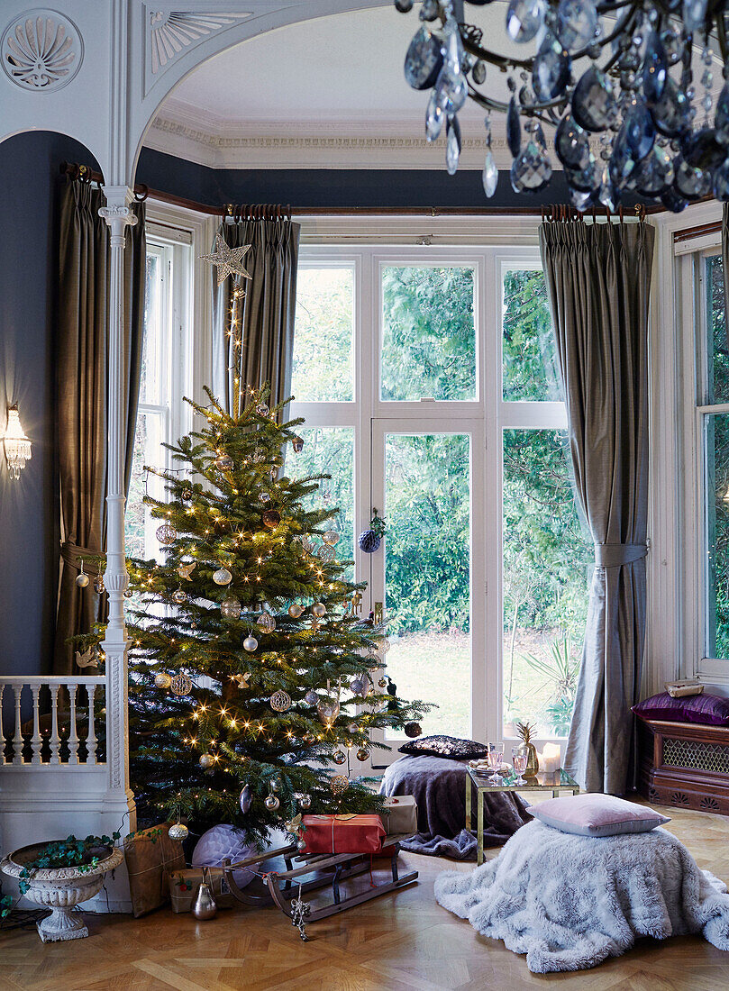 Schlitten unter beleuchtetem Weihnachtsbaum im Erkerbereich eines Hauses in East Grinstead, West Sussex, UK