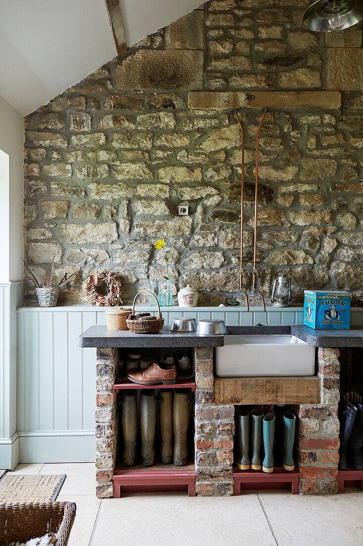 Gummistiefel und Waschbecken mit freiliegender Steinwand im Eingangsbereich eines Bauernhauses in Northumberland, UK