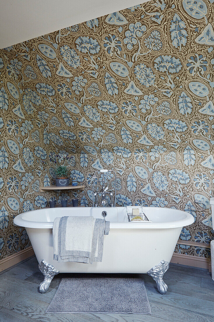 Freestanding clawfoot bath with blue and gold patterned wallpaper in Cotswolds cottage, UK