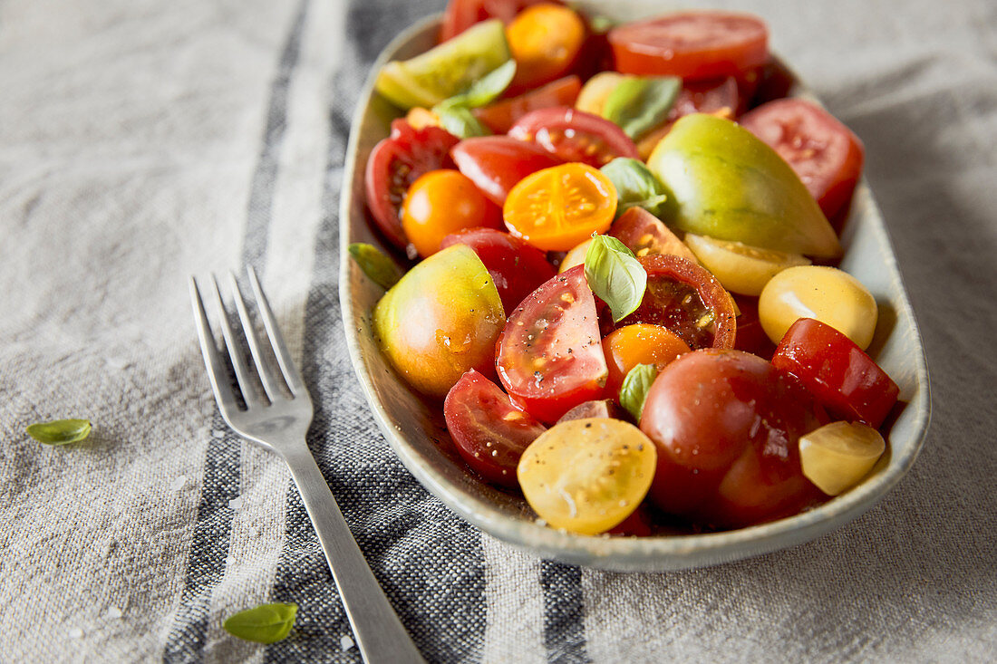 Tomatensalat; bunte Tomaten; Basilikum frisch; Leinentischdecke mit blauem Streifen; kleine Gabel; ovaler Teller
