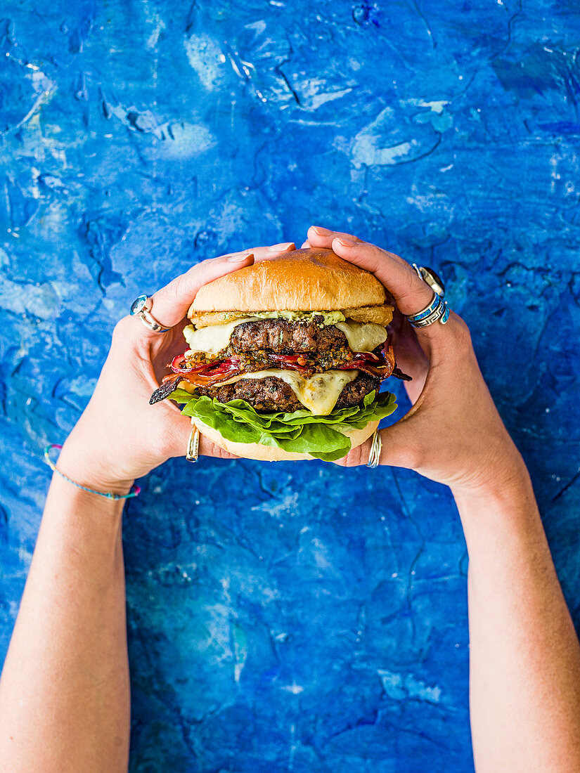 Hände halten brasilianischen Burger mit Käse, Chimichurri-Relish, Peri-Peri-Bacon und Avocado-Mayo