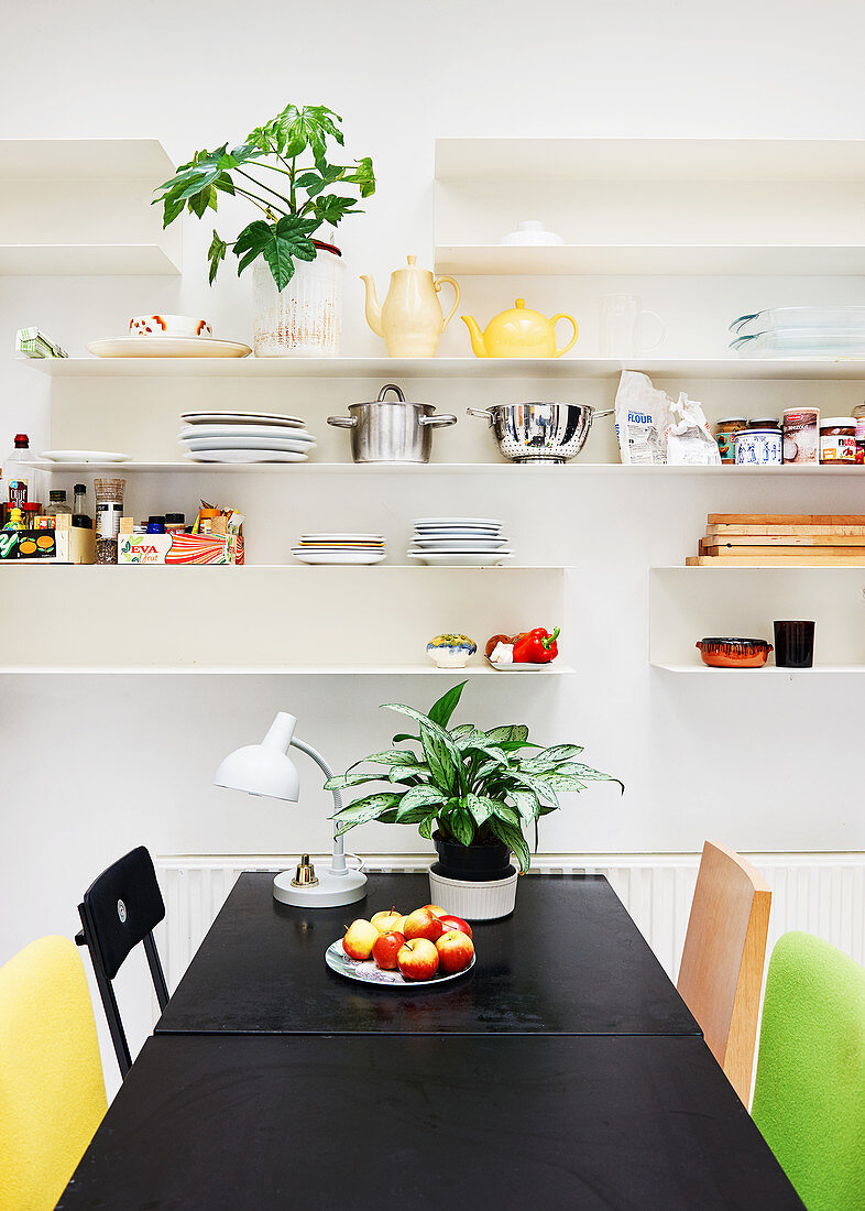 Black table and colourful chairs below crockery on white shelves