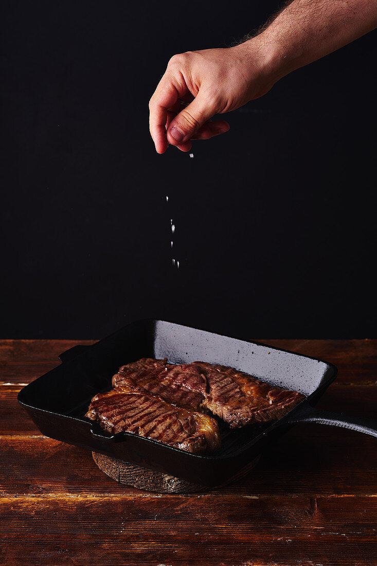 Male hands adding salt to beef steak on grill