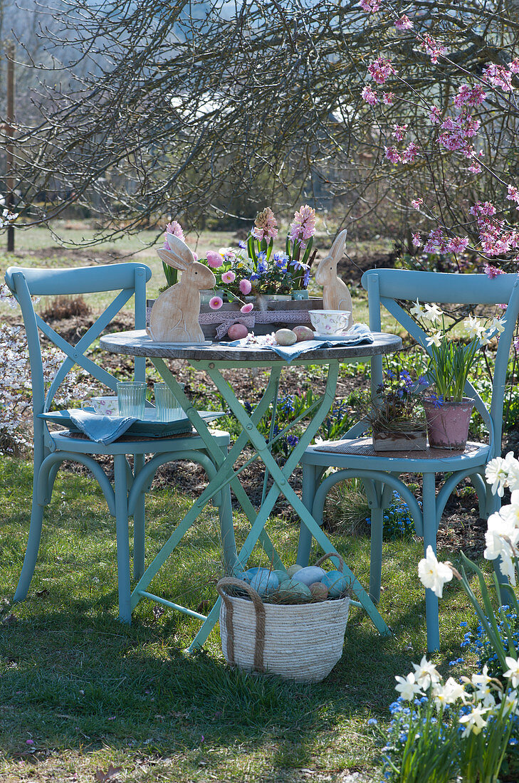 Kleine Sitzgruppe im Frühlingsgarten, Töpfe mit Hyazinthen, Tausendschön, Narzissen 'Toto' und Strahlenanemone, österlich mit Ostereiern und Osterhasen