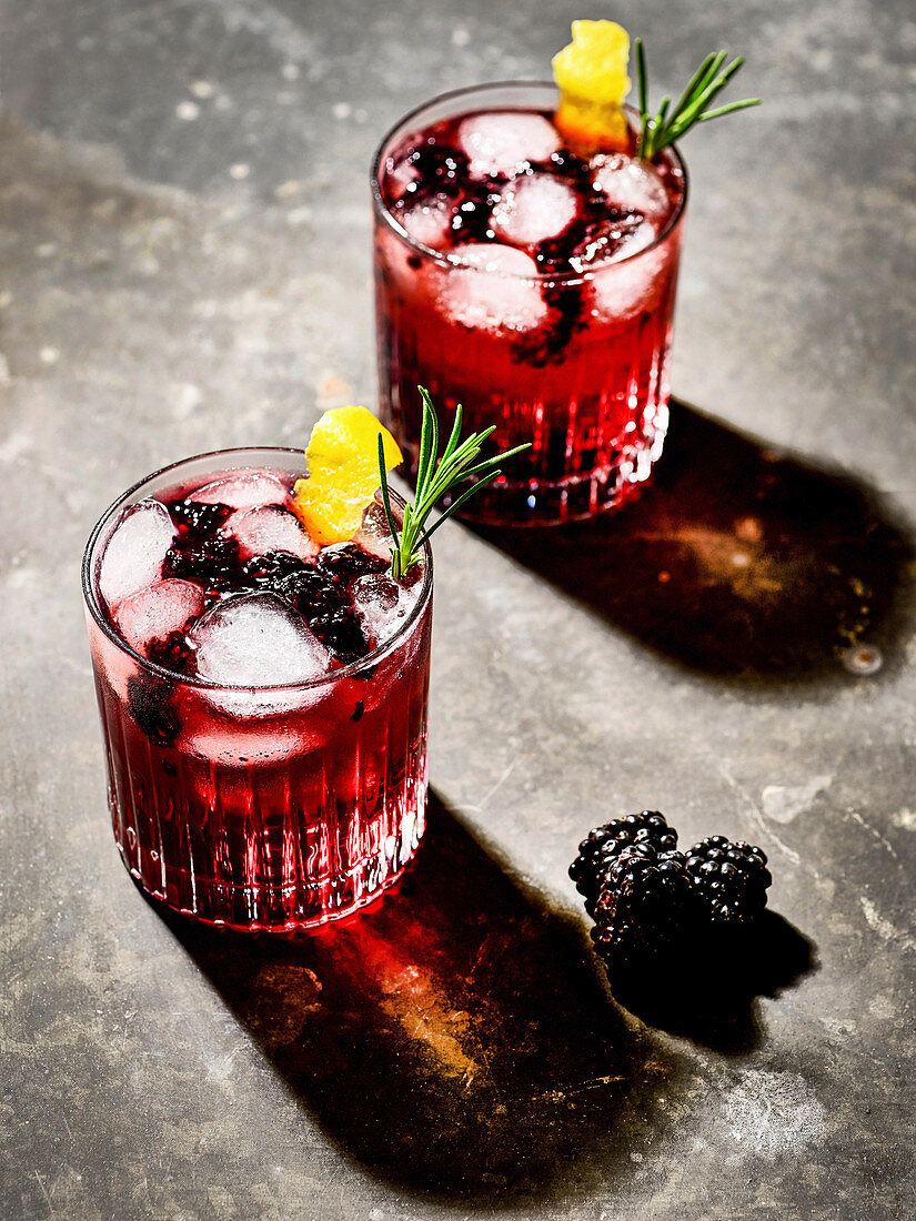 Two glasses of gin and tonic with blackberry juice, blackberries, ice cubes, lemon zest and rosemary