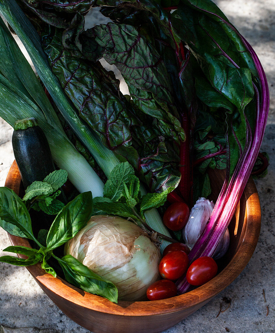 Wooden bowl filled with vegetables and herbs - rainbow swiss chard, leeks, tomatoes, zucchini, onion, garlic, mint and basil