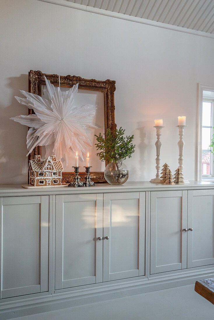 Paper star hung on gilt picture frame and gingerbread house on sideboard