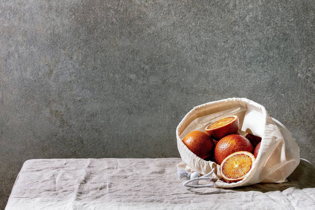 Blood sicilian oranges, ripe and juicy, in cotton eco friendly bag, whole and sliced, on grey linen table cloth, concrete wall as background.