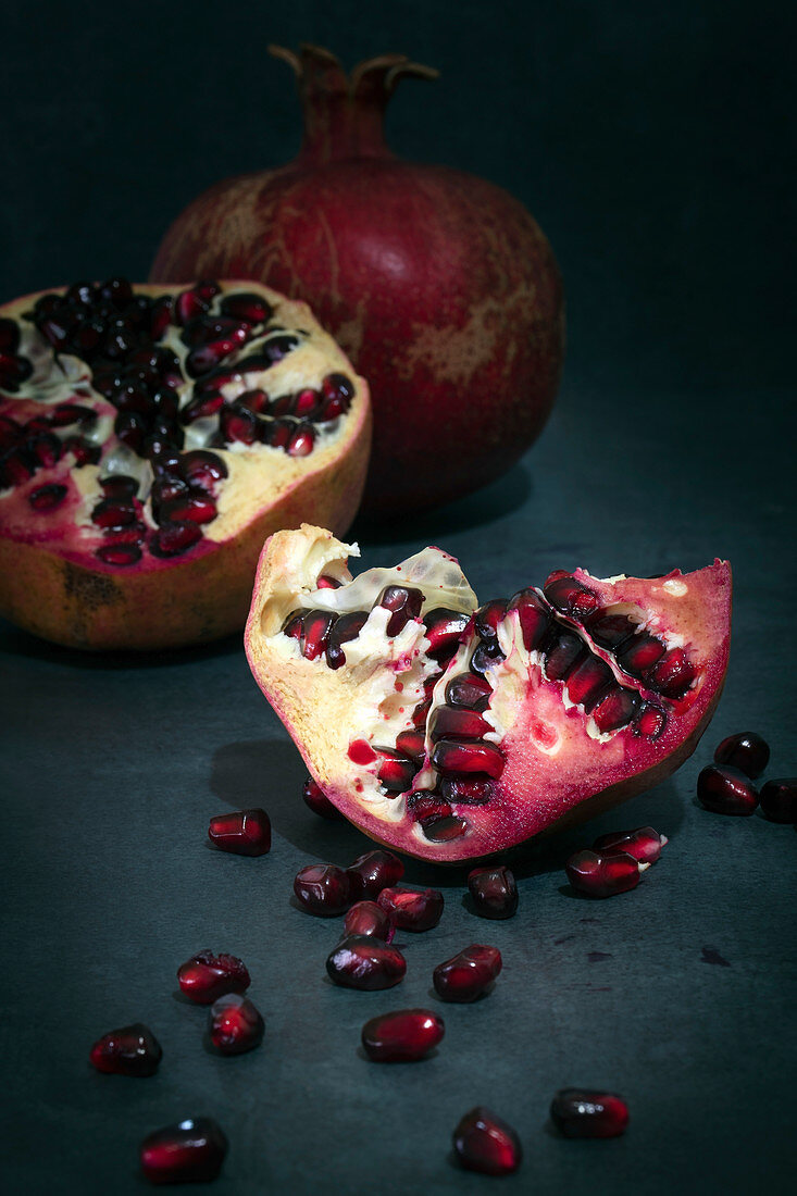 Pomegranate whole and in pieces with scattered seeds on a dark background.